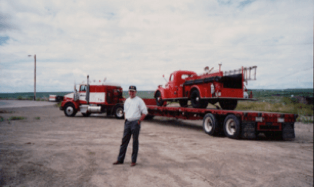 Jim stands in front of a semi-truck and flat-deck trailer with an antique firetruck on it