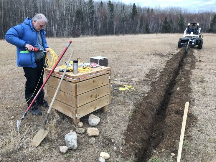 Dr. Martin Connors working in a field while a piece of heavy equipment digs a trench next to him.