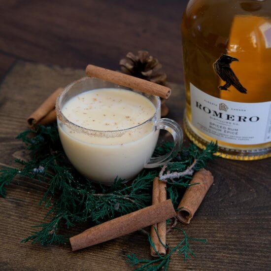 Cup of eggnog with a bottle of rum behind it and cinnamon sticks