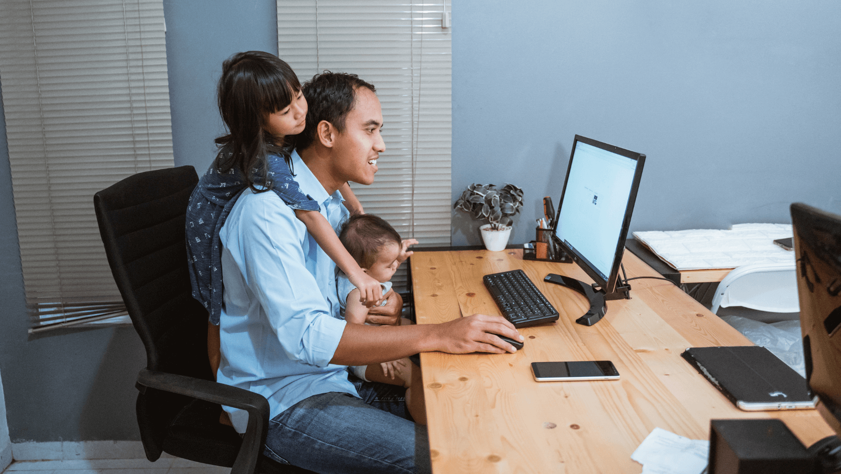 father sitting at computer with a toddler on his shoulders and a baby in his lap