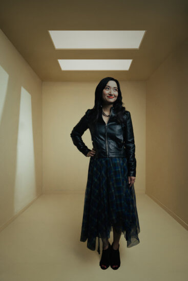 AU MBA grad Laurie Wang standing in an empty room wearing a long black skirt, black shoes and a black leather jacket. She has one hand at her waist.