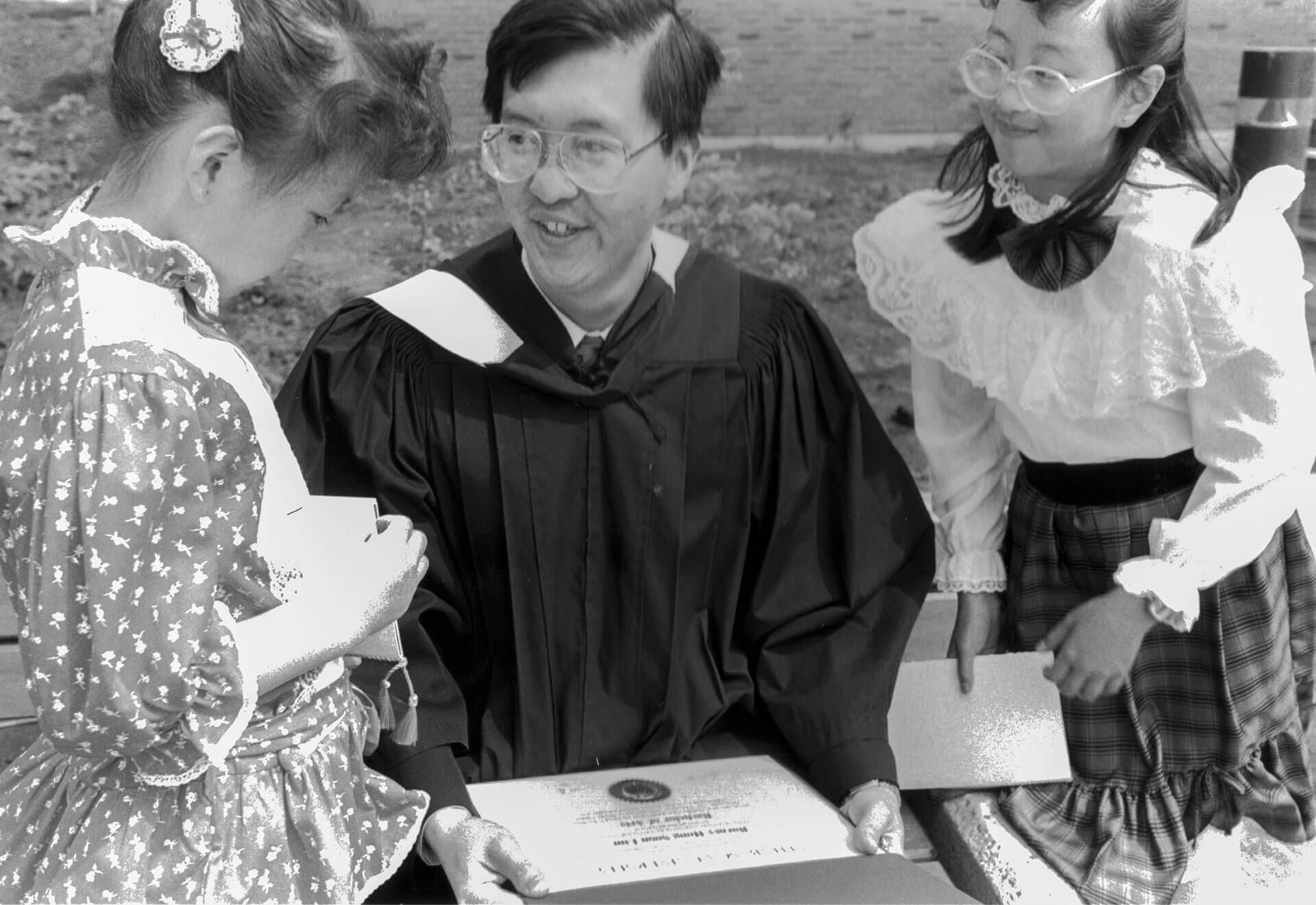 A grad (center) is sitting down holding his parchment, on either side is his two young daughters.