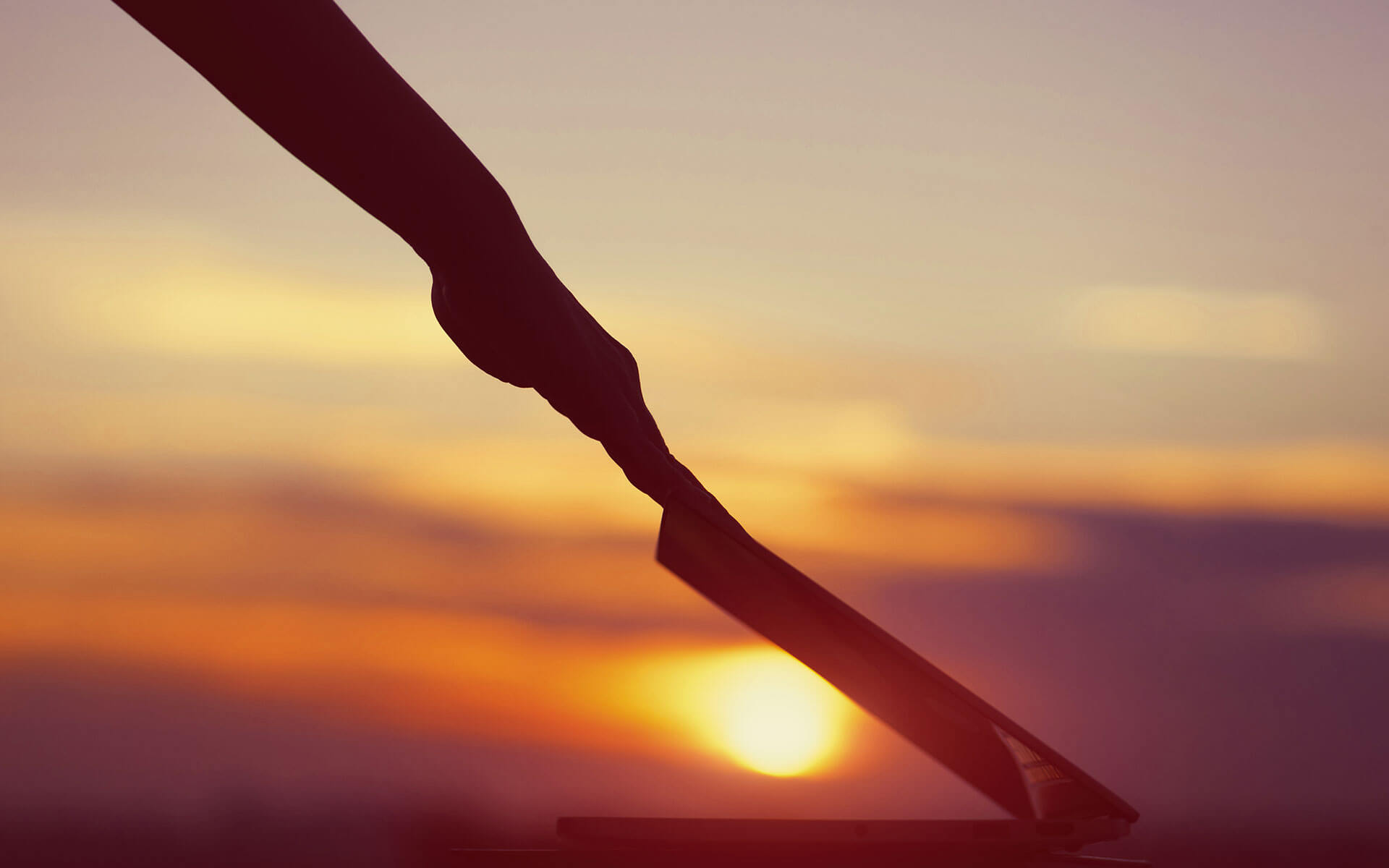 A person's arm extends across the frame to close a laptop with a sunset background